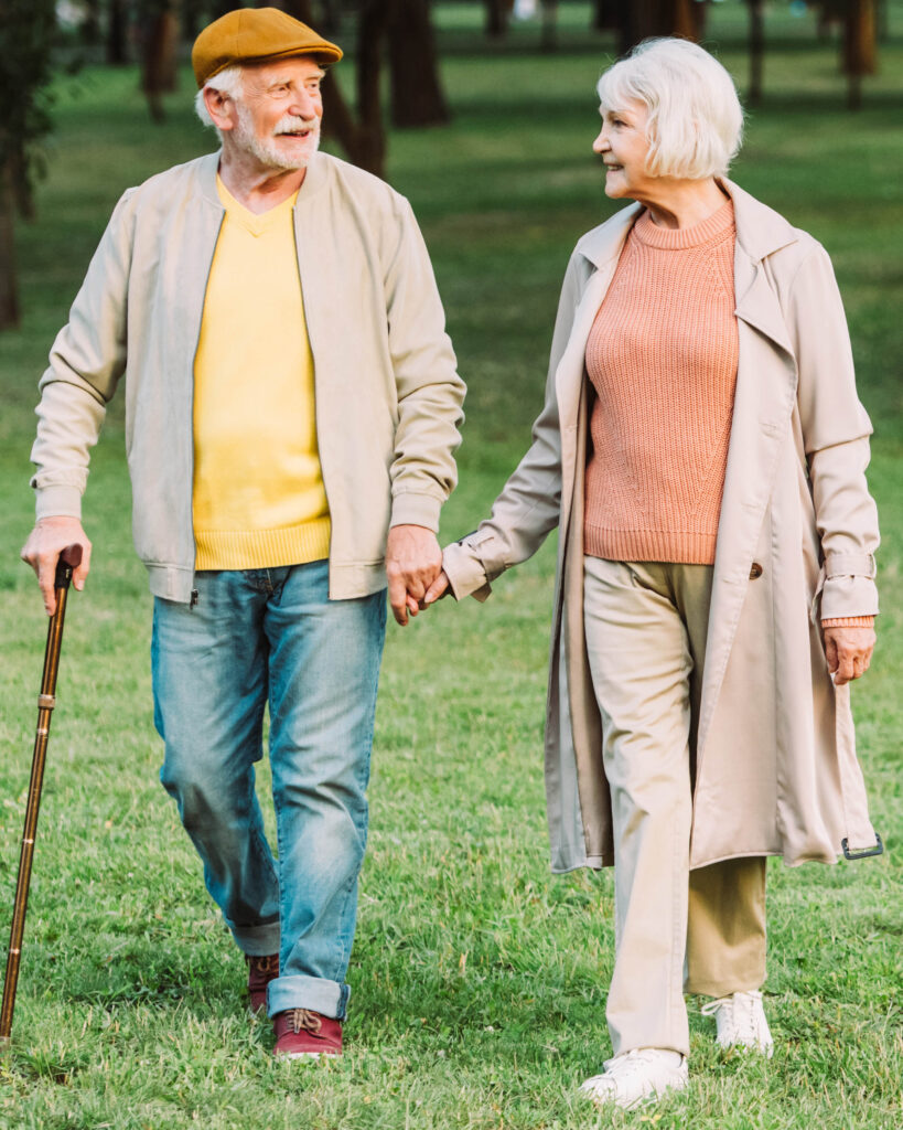 A white-haired senior couple walking hand in hand through a sunlit park, looking lovingly at each other